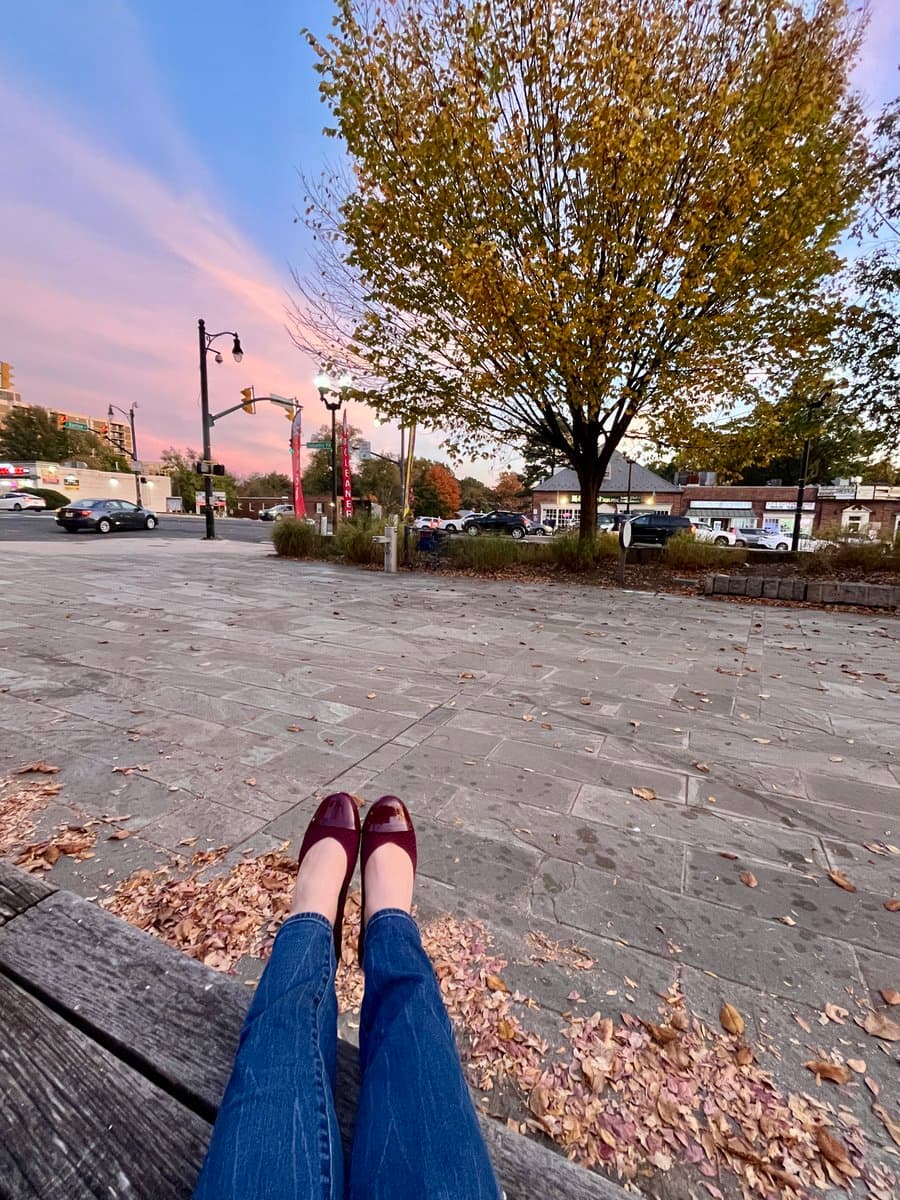 A first-person perspective photo of a person's legs and feet in maroon shoes, resting on a wooden bench in a paved public square. The background features a large tree with autumn leaves, a street with cars, and storefronts under a twilight sky.