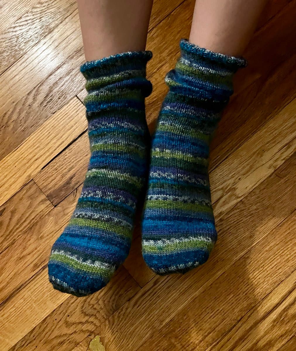 A pair of feet wearing hand-knitted socks with horizontal stripes in shades of blue, green, and white, resting on a wooden floor. The socks have a ribbed cuff and appear to be made from a variegated yarn.