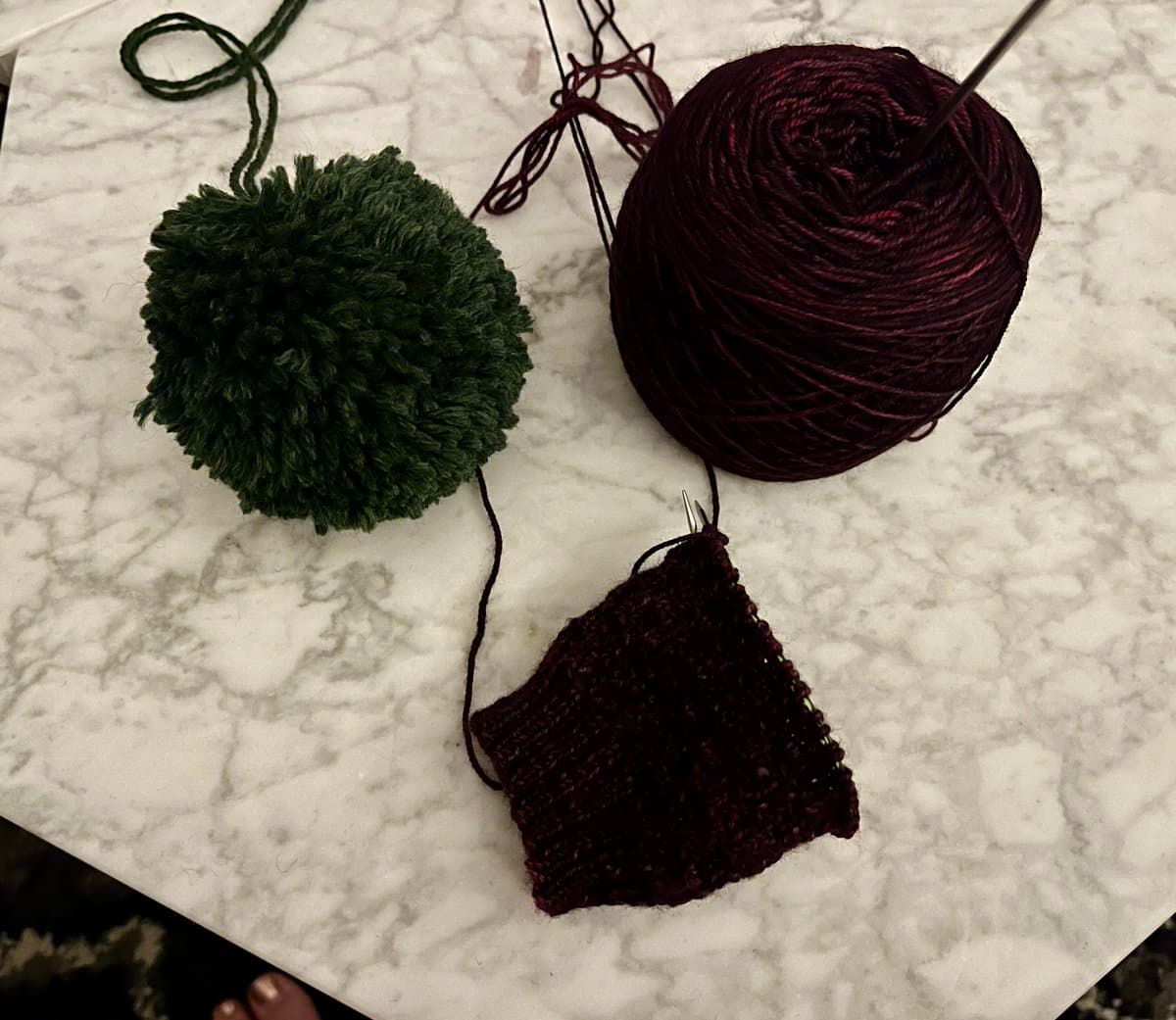 A top-down photo of knitting supplies on a white marble surface, including a dark green yarn pompom, a ball of dark maroon yarn, and a small swatch of maroon knitting on a needle. A person's foot is partially visible at the bottom edge of the frame.