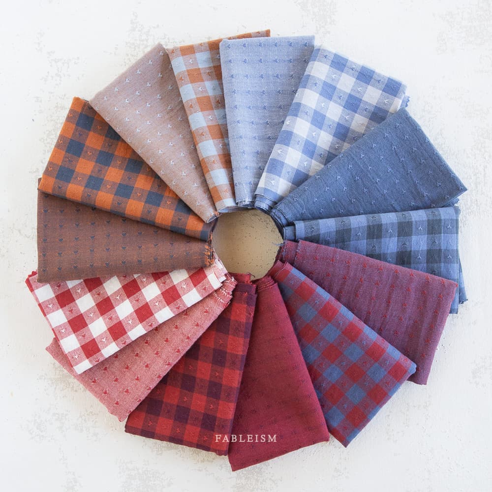 A circular arrangement of folded fabric swatches in various plaid and dotted patterns, featuring autumnal colors like rust, red, and blue. The brand name 'FABLEISM' is printed in white text at the bottom center of the image.