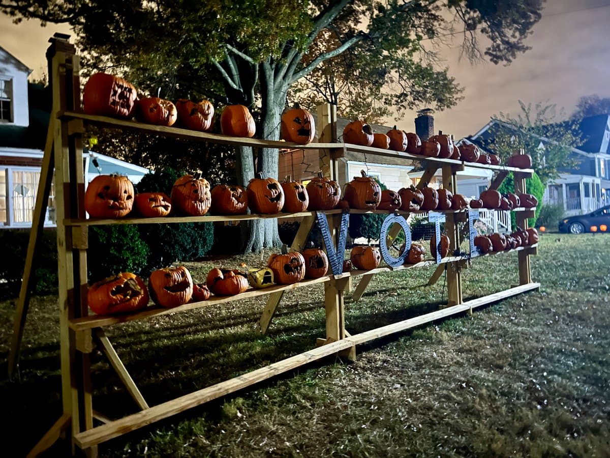 A nighttime photograph of a wooden display rack in a front yard, filled with numerous carved jack-o'-lanterns of various sizes. Large letters spelling "VOTE" are incorporated into the middle shelf of the pumpkin display.