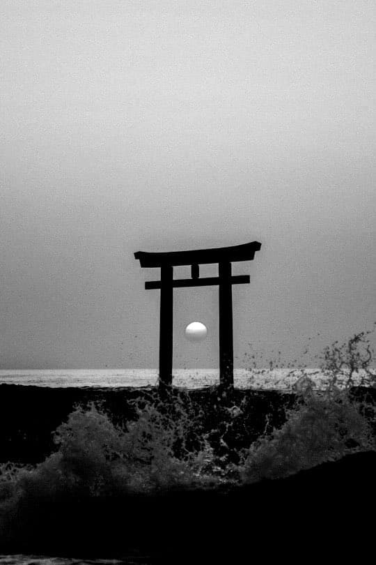 A black-and-white photograph of a Japanese torii gate standing on a rocky shore, with the moon visible through its center and waves crashing in the foreground.