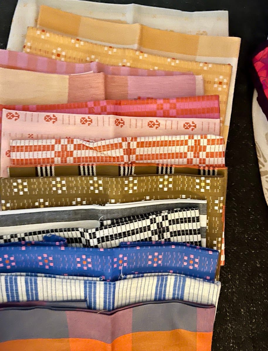 A top-down view of a stack of folded fabric pieces with various woven patterns and colors, including stripes, checks, and geometric motifs. The fabrics are arranged in a fanned-out pile on a dark surface.