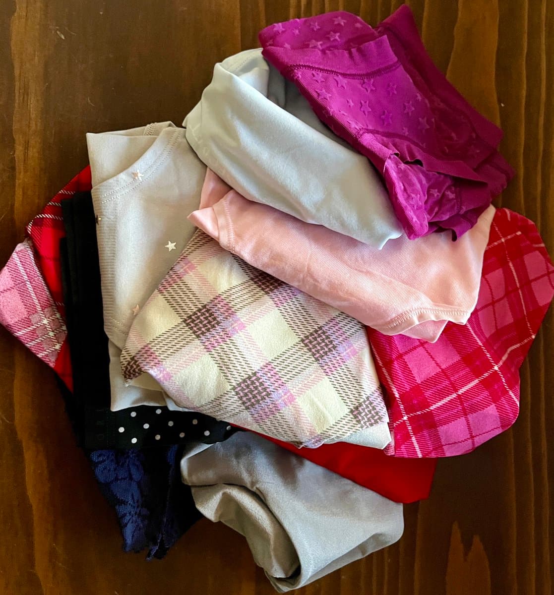 A top-down view of a pile of folded clothing on a wooden floor, featuring various fabrics including a magenta star-patterned top, a cream shirt with small stars, a pink plaid shirt, and a red argyle sweater. The garments are stacked in a casual, colorful heap.