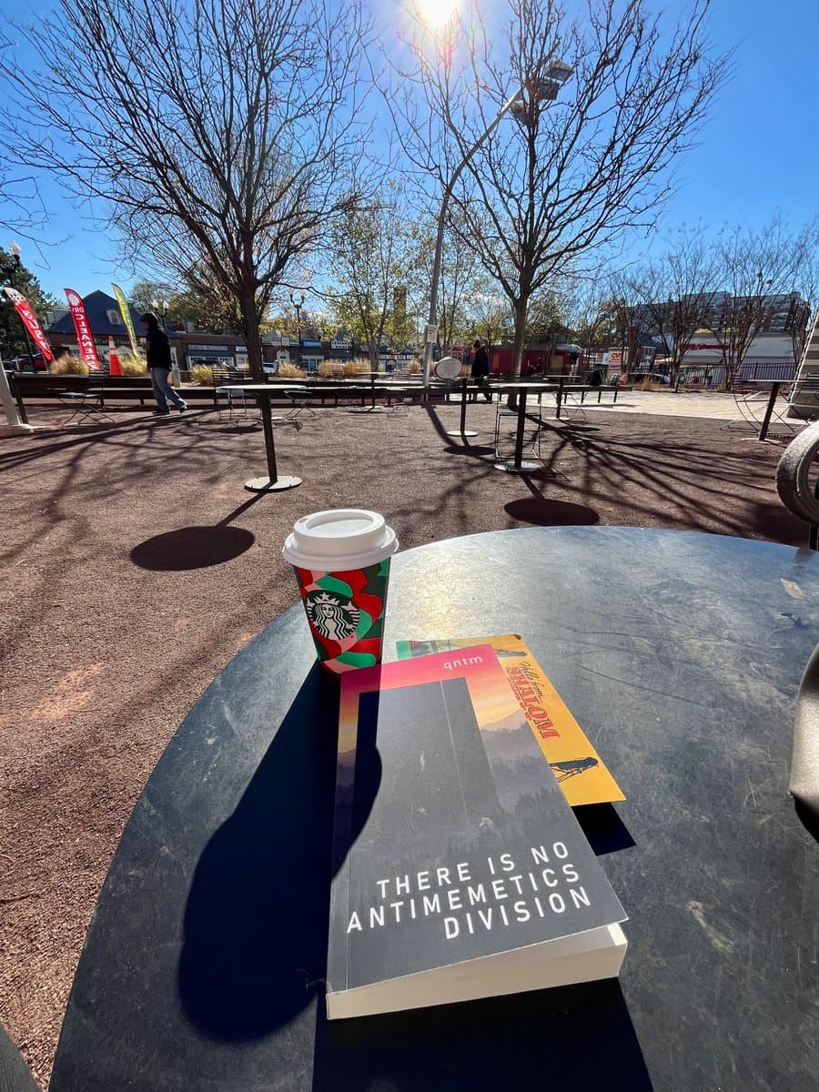 A first-person view of a black outdoor table holding a Starbucks coffee cup and two books, with a park setting in the background. The top book has a dark cover with white text, while the book underneath has a yellow and orange cover. Bare trees, picnic tables, and a few people are visible in the sunny, open area behind the table.