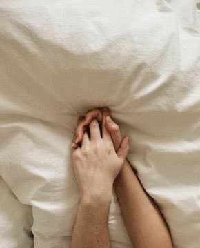 A close-up photograph of two hands gently clasped together, resting on a soft, white bed sheet or duvet. The image conveys a sense of intimacy and tenderness through the simple gesture of holding hands.
