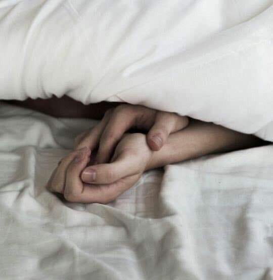 A close-up photograph showing two hands gently intertwined under a white blanket on a bed. The image conveys a sense of intimacy and connection between the two people.