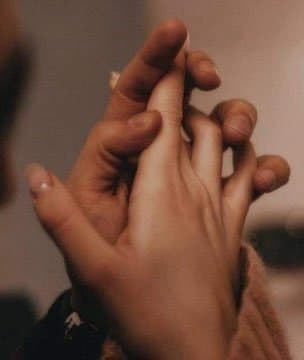 A close-up photograph showing two pairs of hands gently intertwined, with fingers interlaced in a gesture of intimacy or connection. The image has a warm, soft-focus quality with a shallow depth of field that blurs the background.
