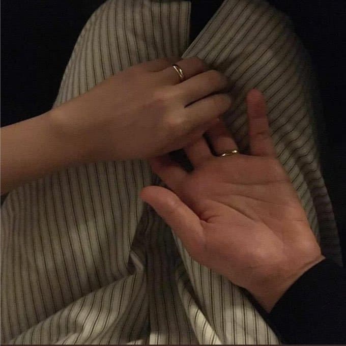 A close-up photograph of two hands gently touching over a person's lap, which is covered in striped fabric. One hand rests on top of the other, and both are wearing simple gold rings.