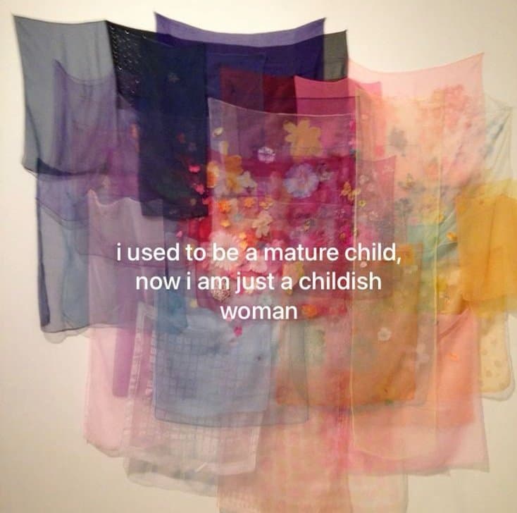 A photograph of a colorful, abstract textile art piece made of layered, translucent fabrics in shades of blue, purple, pink, and orange, with white text overlaid in the center.