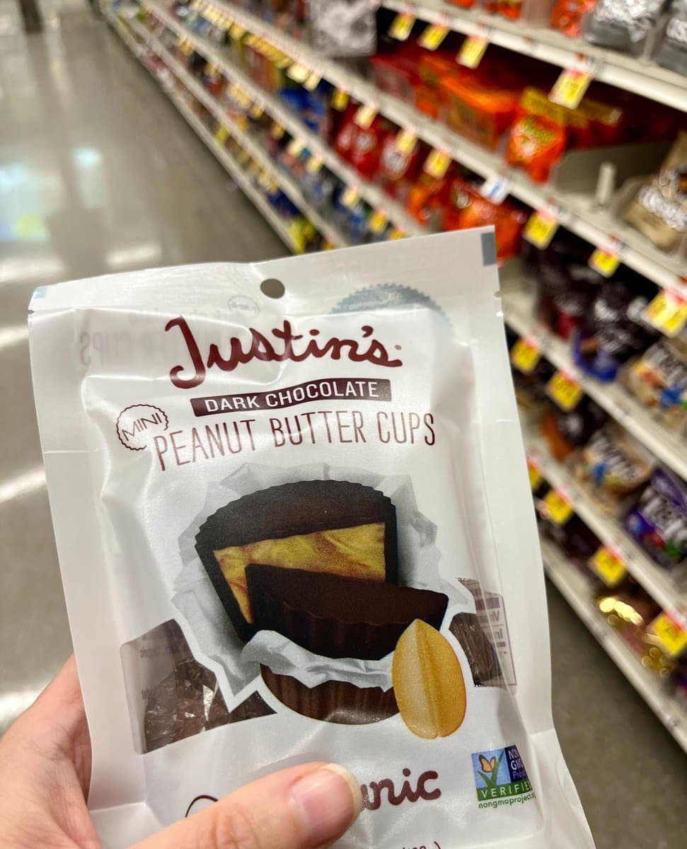 A close-up photograph of a hand holding a package of Justin's Mini Dark Chocolate Peanut Butter Cups in a grocery store aisle.