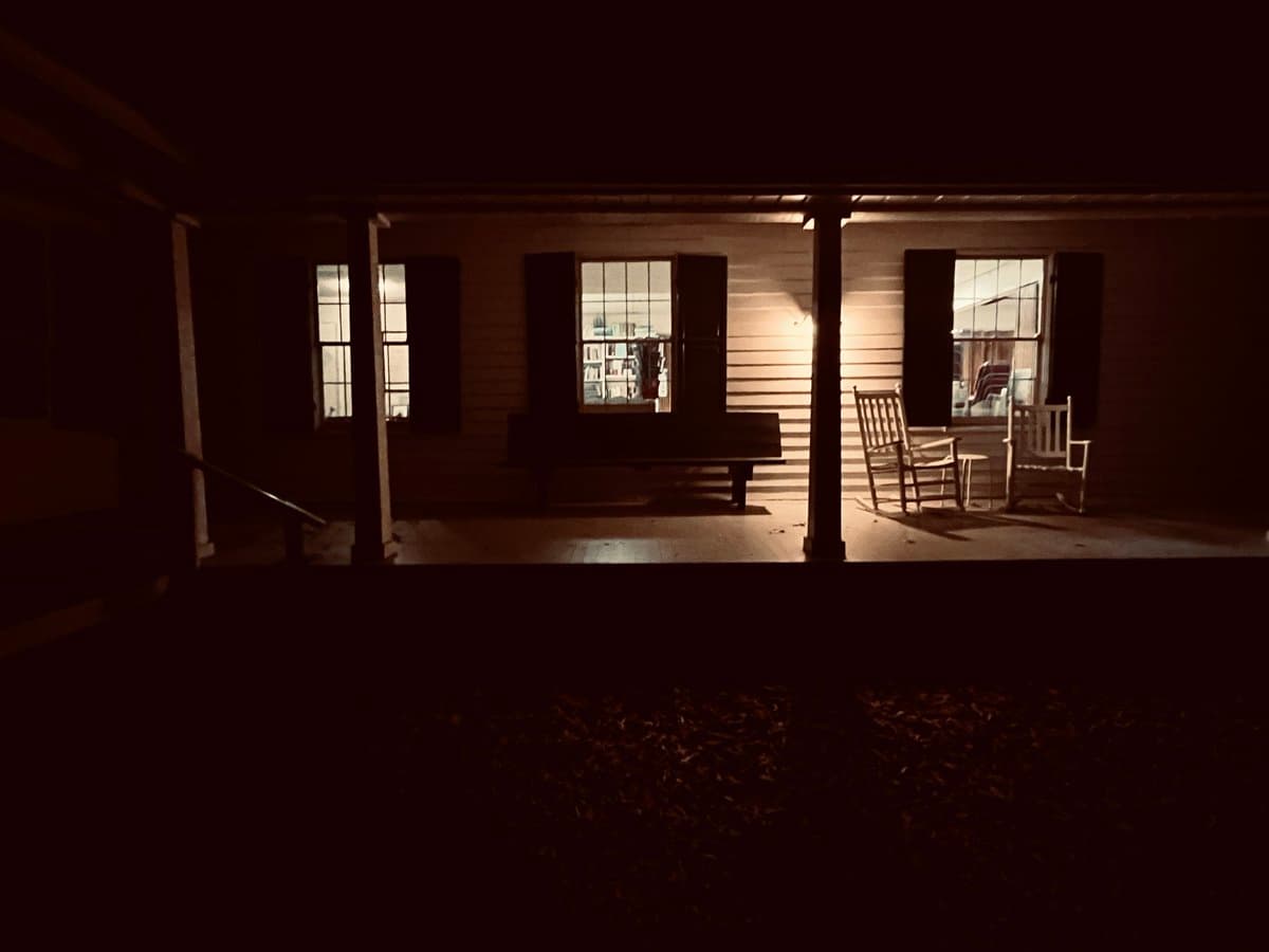 A nighttime photograph of a house's front porch, illuminated by light spilling from the windows. The porch features a wooden bench and two rocking chairs, with the surrounding area in deep shadow.