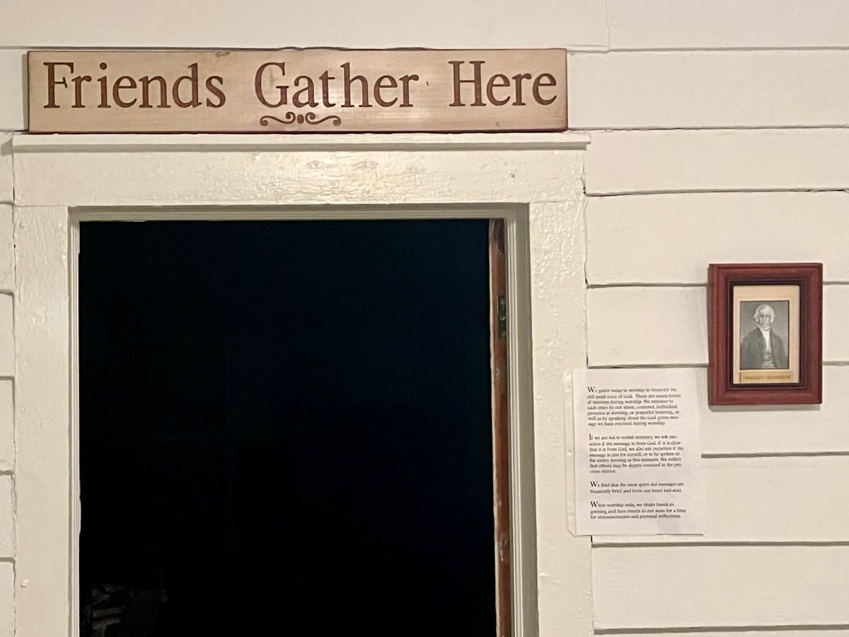 A photograph of a white-painted wooden doorway with a dark interior, topped by a wooden sign reading "Friends Gather Here." To the right of the door, a framed portrait of a man and a printed sheet of text are mounted on the wall.