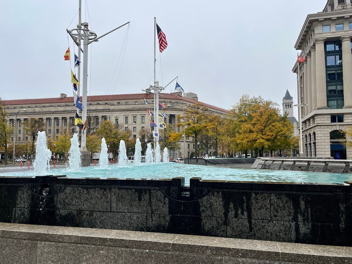 A photograph of a city plaza featuring a large, rectangular fountain with multiple water jets in the foreground. In the background, there is a large, classical-style government building with a red roof, surrounded by trees with autumn foliage and tall flagpoles flying various national flags, including the United States flag.