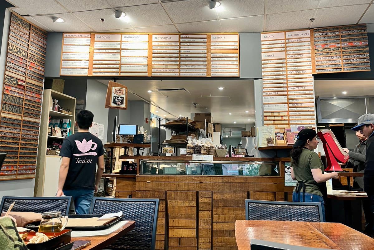 A wide interior shot of a cafe or bakery featuring a wooden service counter, a glass display case, and a large menu board mounted on the wall above. A customer in a black 'Team' t-shirt stands at the counter while a staff member in a green shirt works behind it.