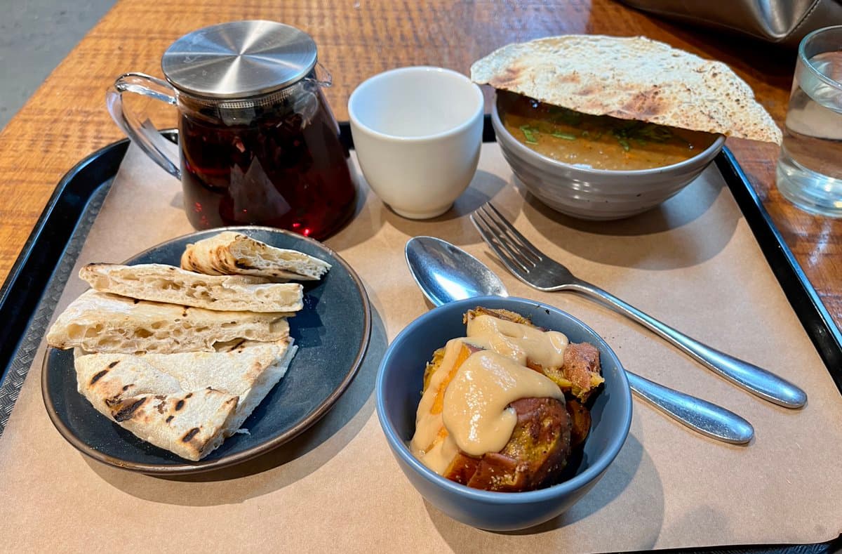 A meal served on a black tray, featuring a glass teapot with dark tea, a white cup, a bowl of soup with a piece of flatbread, a plate of sliced flatbread, and a bowl of food topped with a creamy sauce.