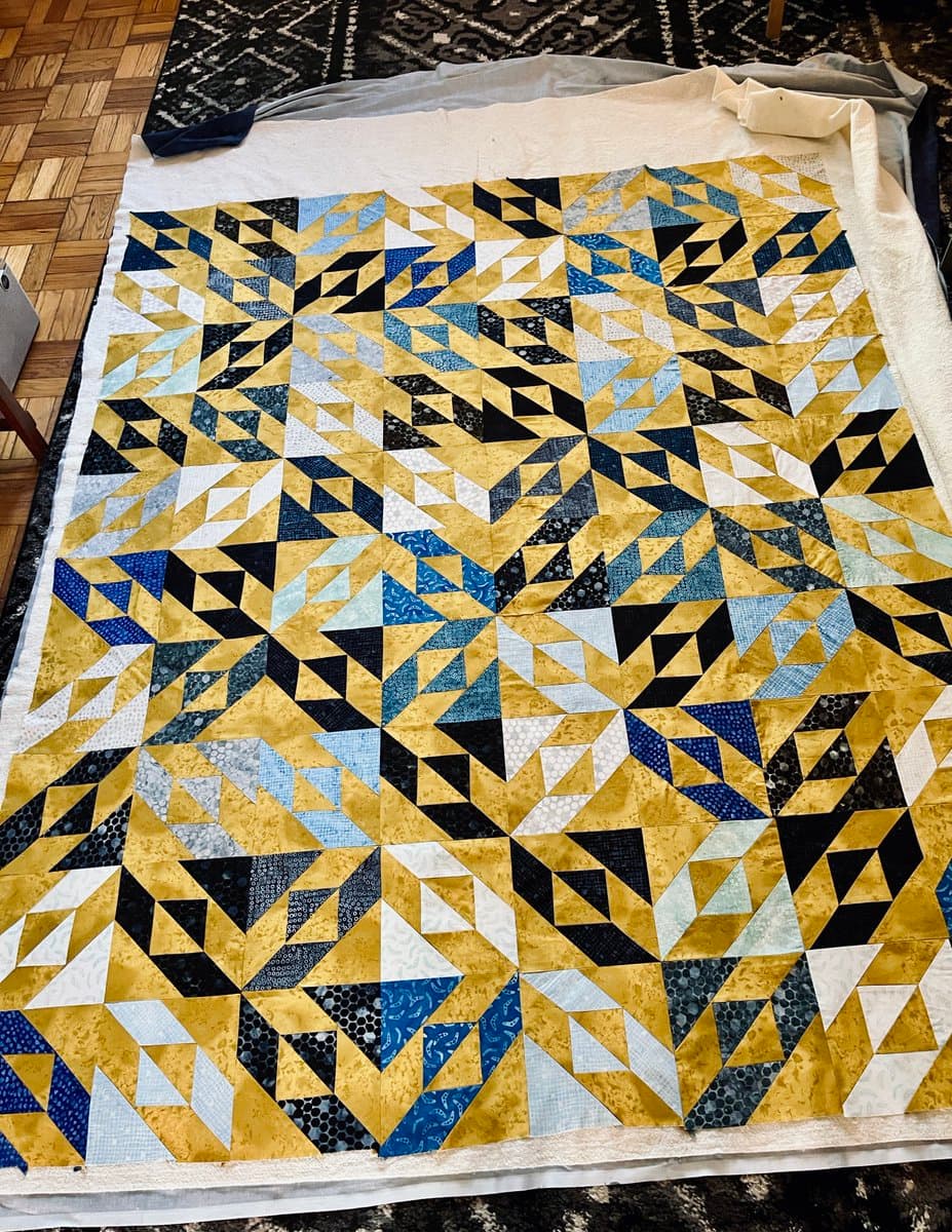 A large, in-progress quilt is laid flat on a floor, featuring a complex geometric pattern of triangles in shades of blue, black, white, and gold. The quilt top is bordered by a plain white fabric, and the scene is set on a wooden floor with a dark rug visible in the background.