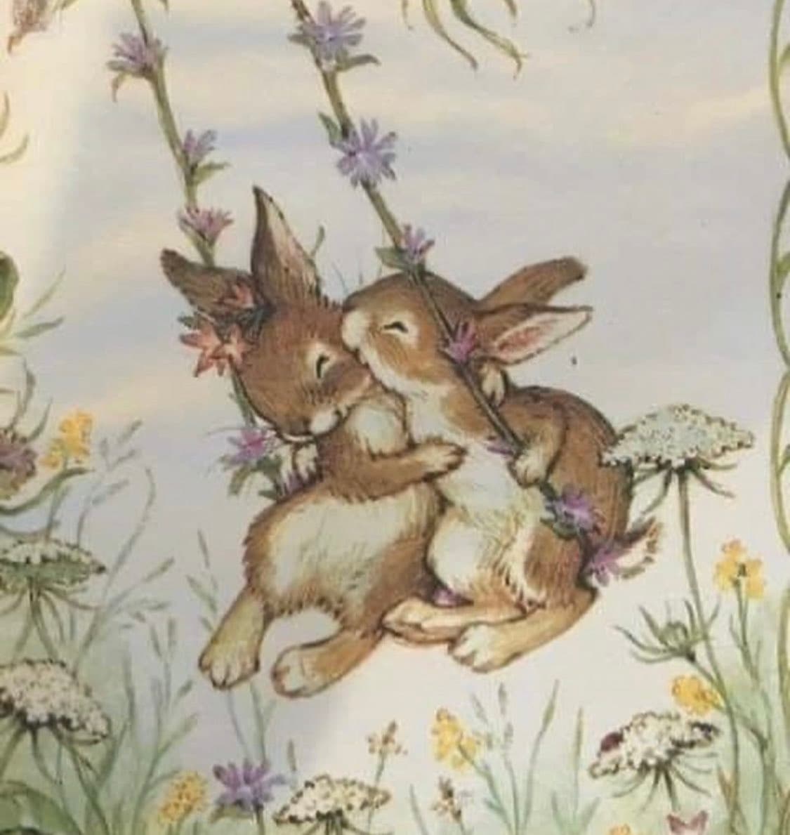 A watercolor illustration depicts two brown and white rabbits embracing each other while hanging from a flowering vine. They are surrounded by various wildflowers, including purple and yellow blooms, against a soft, light background.