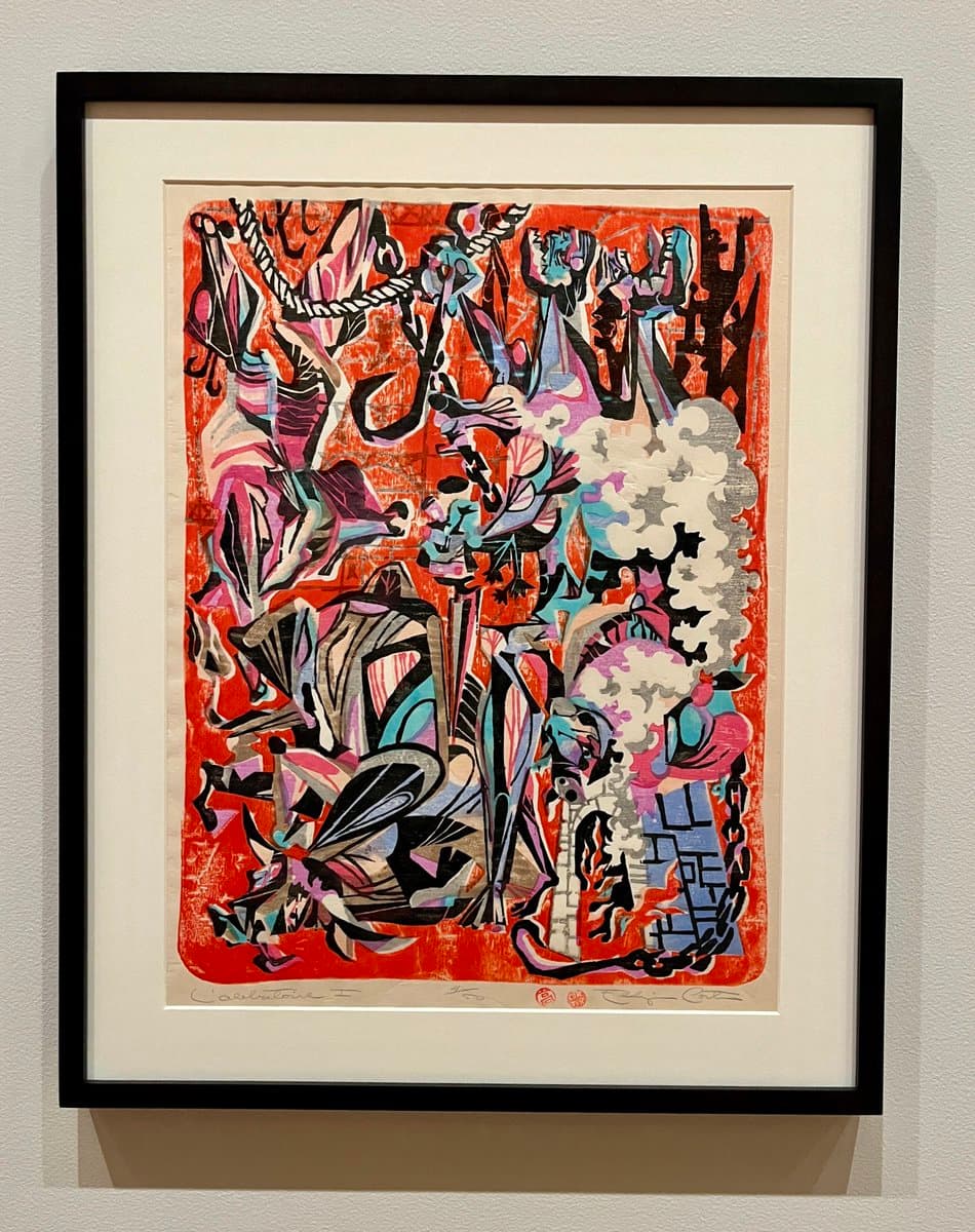 A framed piece of Haitian art hanging on a white wall, featuring a vibrant, abstract composition with bold red, black, and purple colors and expressive, gestural figures. The artwork is matted in white and set in a simple black frame.