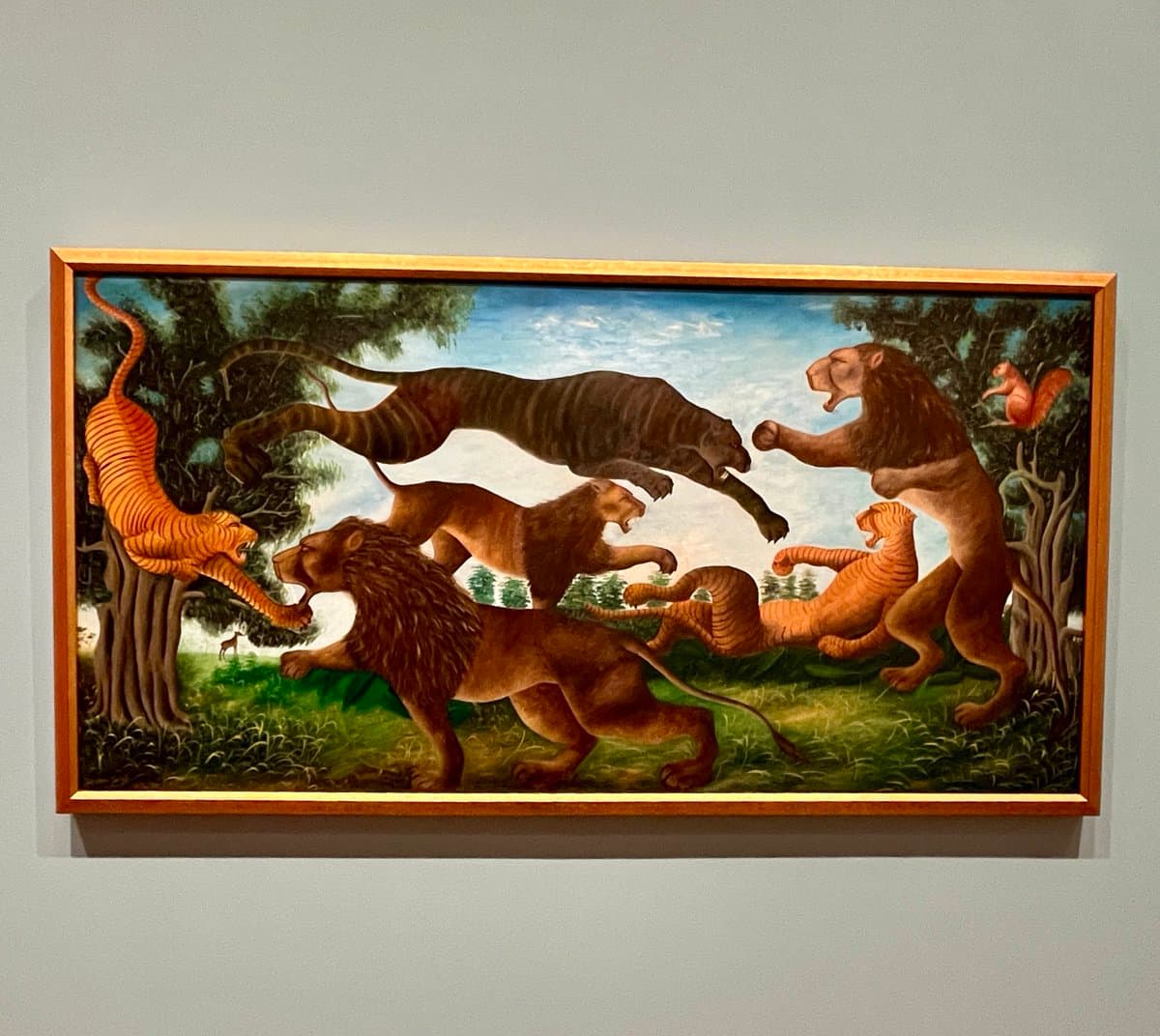 A framed painting hanging on a gallery wall depicts a dynamic scene of multiple large felines, including lions and tigers, interacting in a grassy landscape with trees. The artwork is presented in a simple wooden frame.