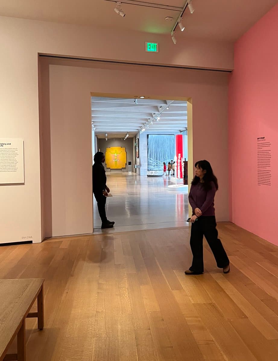 A photograph of a museum gallery with light wood floors and white and pink walls, showing two visitors standing in the foreground and a view into a larger exhibition hall in the background. The space features track lighting, an illuminated green exit sign, and informational placards on the walls.