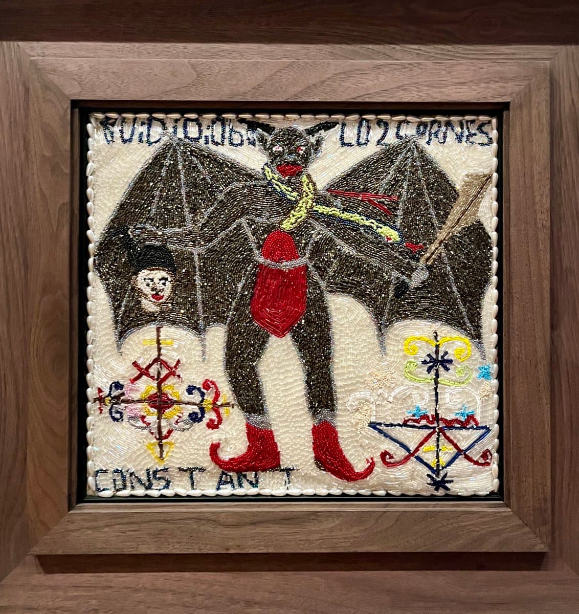 A framed piece of Haitian art, likely a taptap or beadwork piece, depicting a dark, winged figure with red boots and a red loincloth. The figure holds a staff and a small doll, surrounded by colorful, symbolic patterns and text.