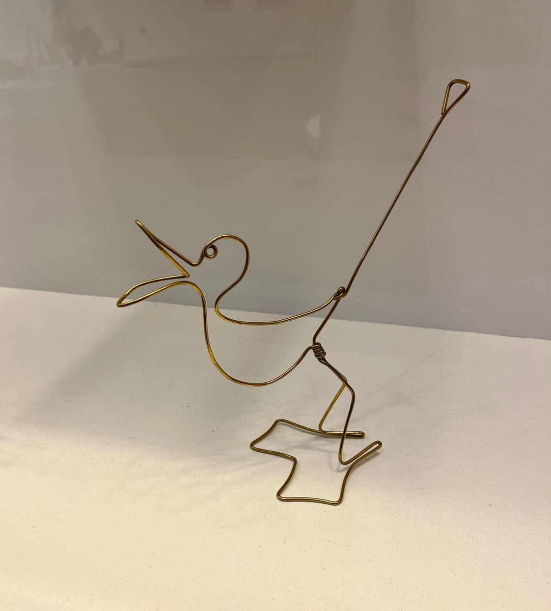 A gold-colored wire sculpture of a bird with its beak open, perched on a star-shaped base. The sculpture is positioned on a white surface against a plain, light-colored wall.