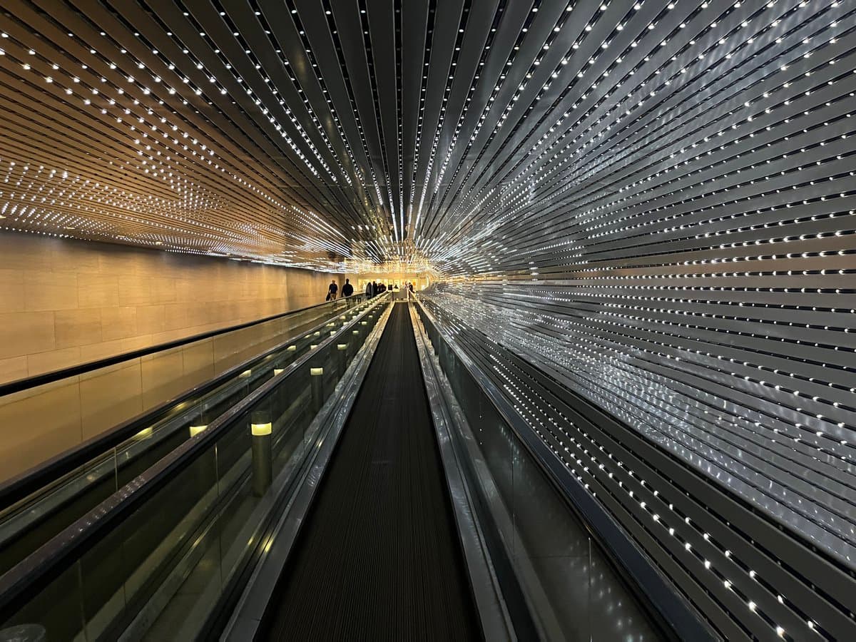 A wide-angle photograph of a long, modern moving walkway tunnel featuring a curved ceiling and walls lined with slats and numerous small lights. The perspective leads toward a bright opening in the distance where a few people are visible.