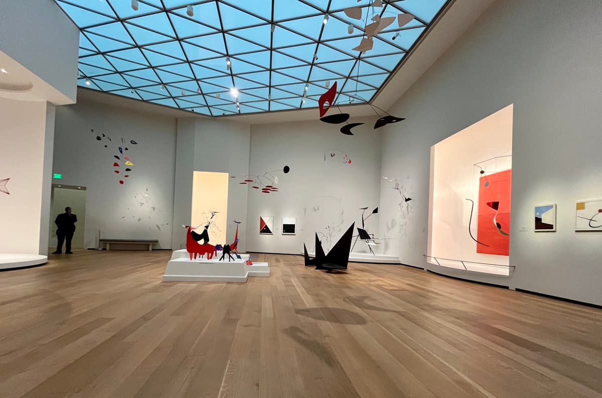 A wide-angle photograph of a bright, modern art gallery featuring a large glass skylight ceiling, light wood flooring, and white walls displaying abstract paintings and suspended mobile sculptures. A person stands near a doorway on the left side of the room.