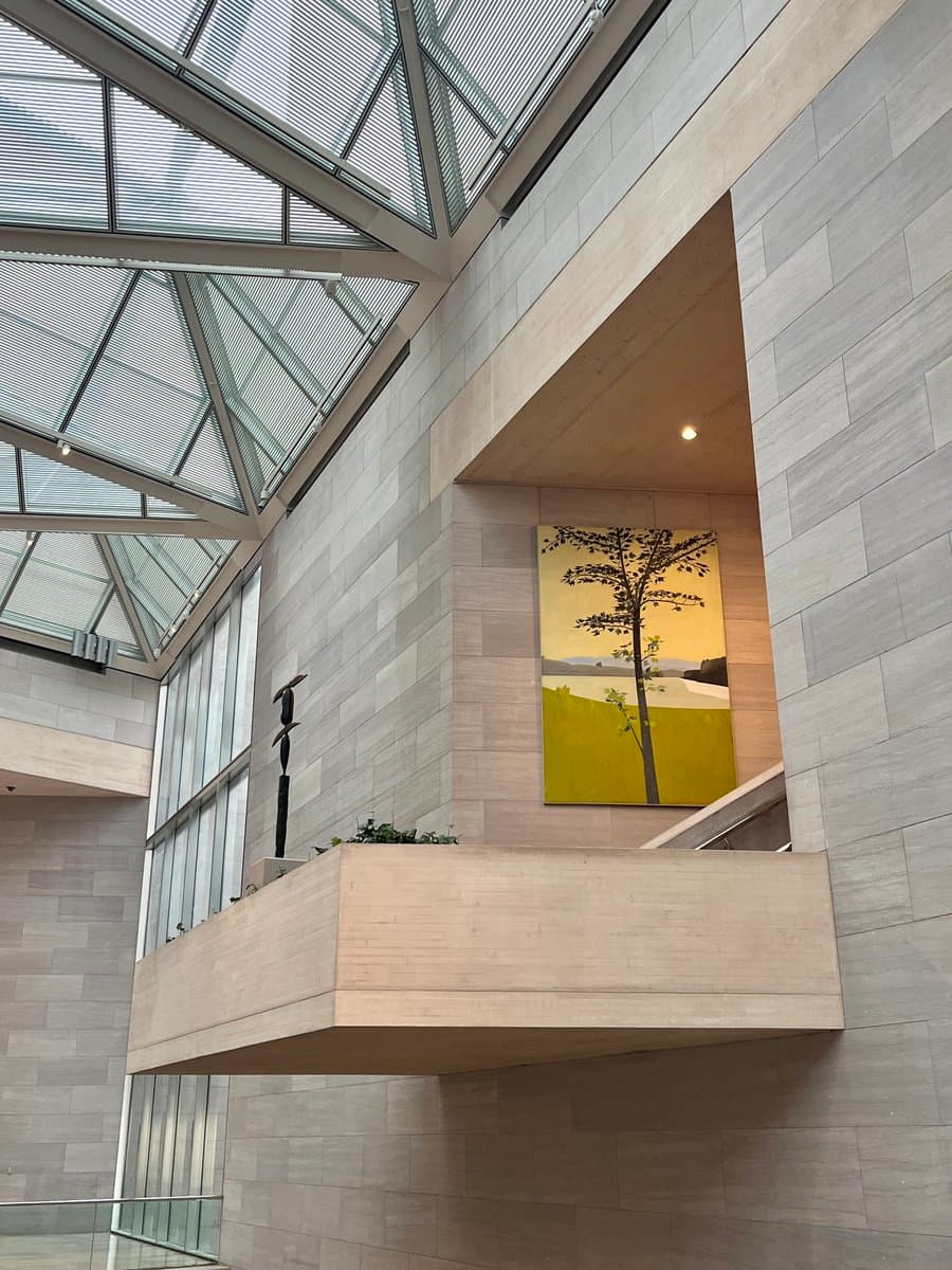 A low-angle interior photograph of a modern building featuring a large, geometric glass skylight and light stone walls. A cantilevered wooden balcony holds a dark sculpture, while a large painting of a tree against a yellow background hangs on the wall in a recessed alcove.