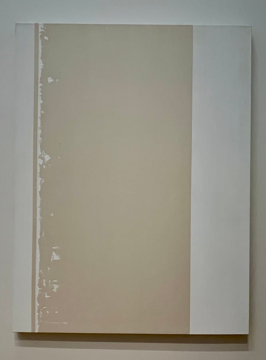 A minimalist abstract painting featuring a large, solid beige rectangular block on a white canvas, with a textured, uneven edge on the left side. The artwork is displayed on a plain wall, emphasizing its simple geometric composition and neutral color palette.