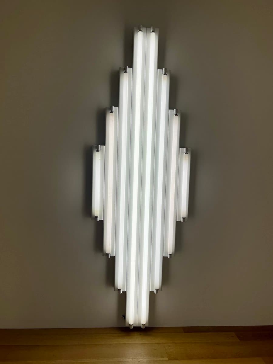 A minimalist wall-mounted light fixture featuring a symmetrical arrangement of vertical fluorescent tubes of varying lengths against a plain grey wall.