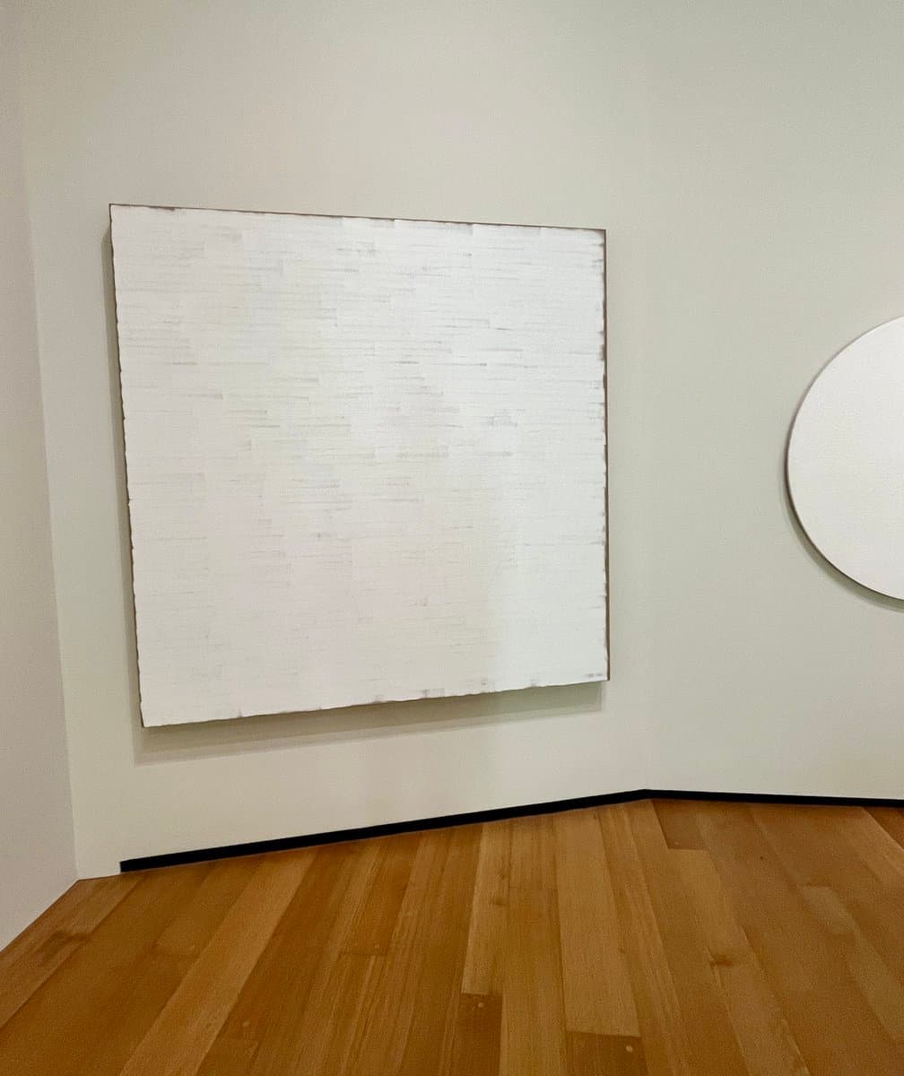 A minimalist gallery corner featuring a large, textured white canvas on the left wall and a partial view of a circular white artwork on the right wall, set against plain white walls and a wooden floor.
