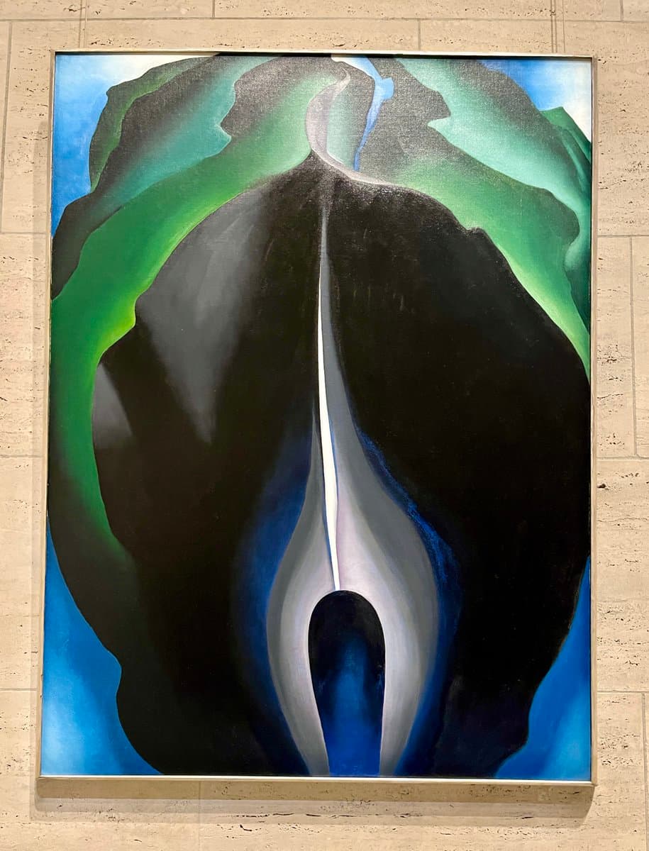 A framed abstract painting hanging on a light-colored stone wall, featuring large, organic shapes in black, green, and blue.