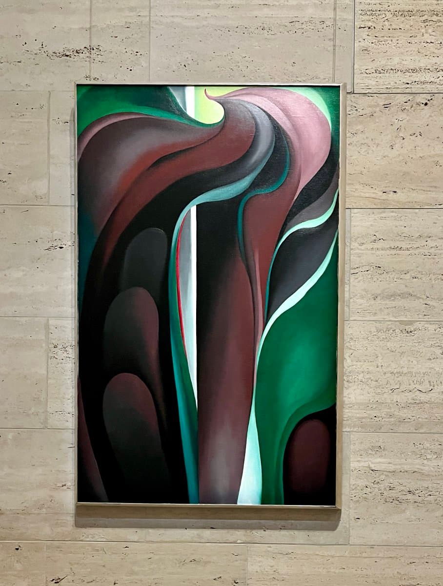 A framed abstract painting featuring flowing, organic shapes in shades of green, brown, black, and white hangs on a textured, light-colored stone wall.