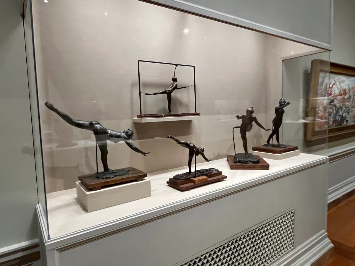 A photograph of a museum display case containing five bronze sculptures of a female gymnast in various poses, including a handstand and a split. The sculptures are mounted on wooden bases within a glass case, with a framed painting visible in the background.