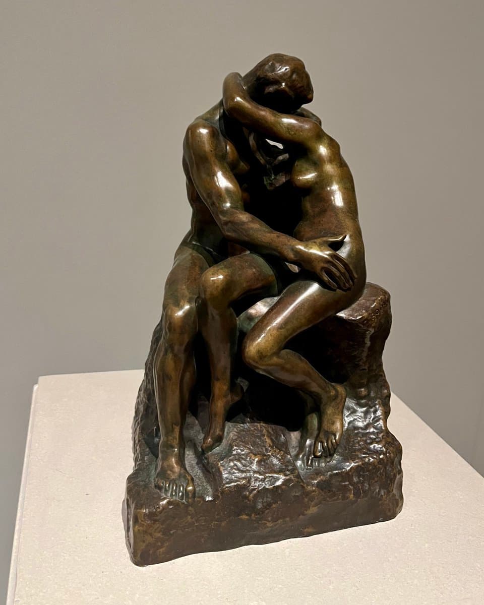 A bronze sculpture of two intertwined, seated figures on a rocky base, displayed on a white pedestal against a plain gray wall. The figures are shown in a close embrace, with one figure's arm wrapped around the other.