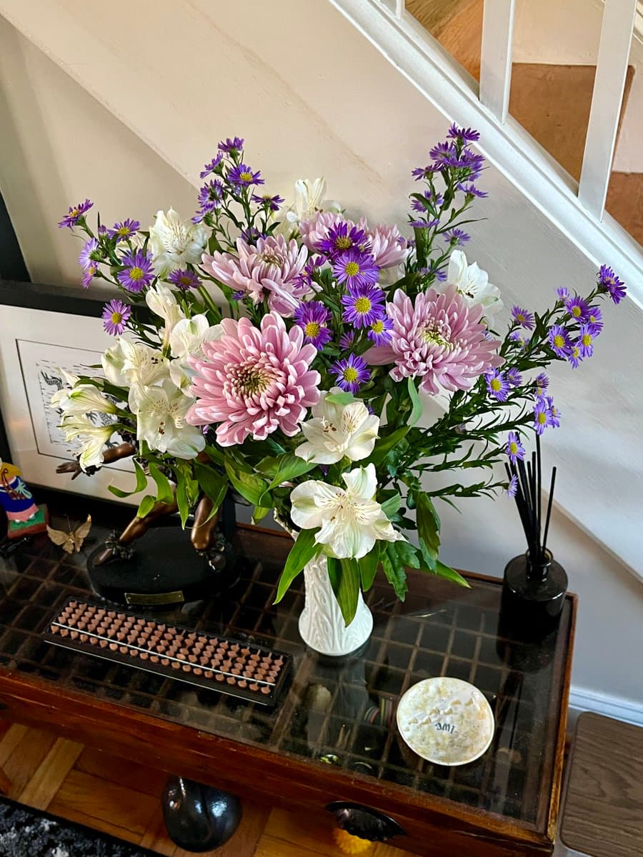 A photograph of a vibrant flower arrangement in a white vase, featuring pink chrysanthemums, white alstroemeria, and purple asters, sitting on a glass-topped wooden table next to a framed picture and a small sculpture.