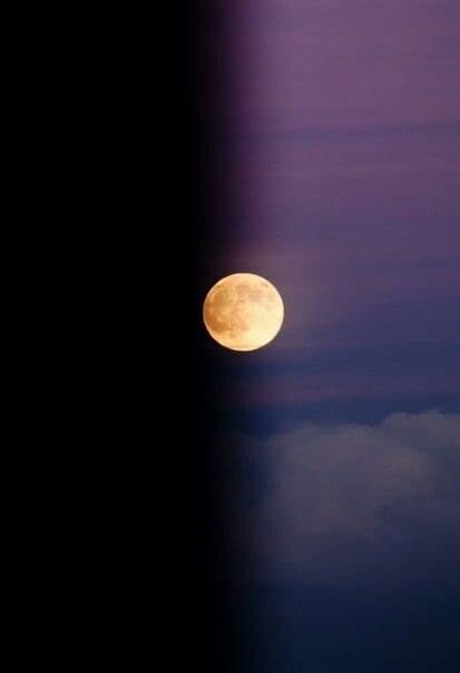 A full moon glowing in a twilight sky with purple and blue hues, partially obscured by a dark vertical edge on the left side of the frame. Wispy clouds are visible below the moon.