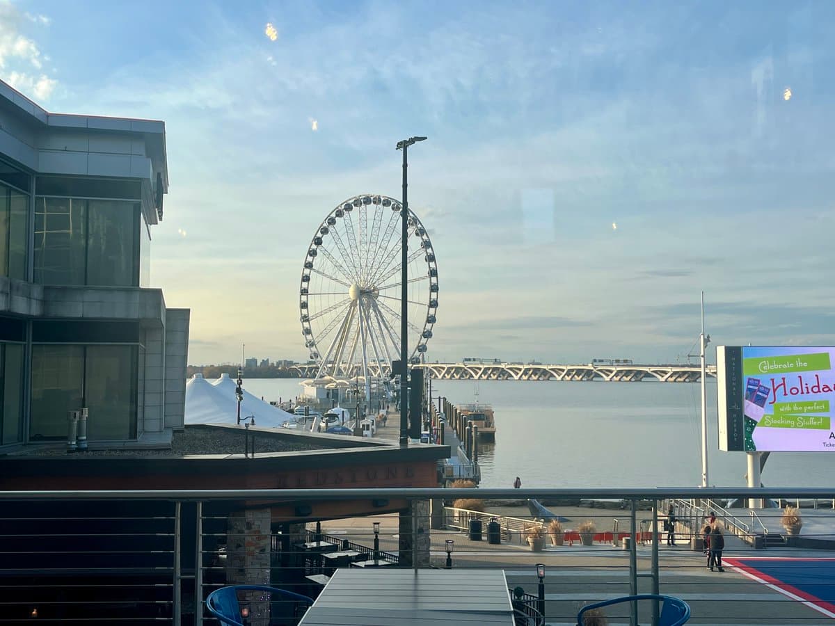 A scenic view of a waterfront promenade featuring a large Ferris wheel and a bridge in the distance under a partly cloudy sky. In the foreground, there is a modern building on the left and a digital billboard on the right.