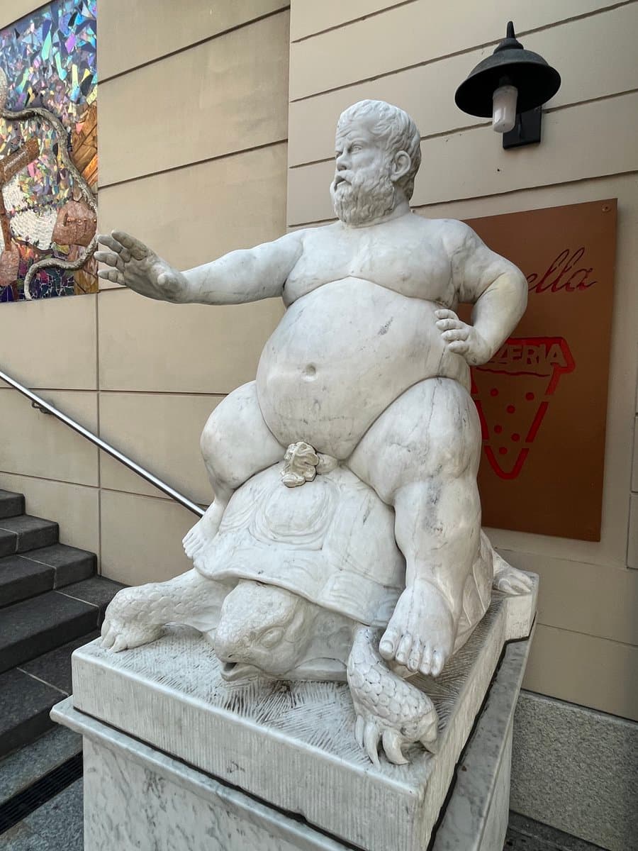 A photograph of a white marble statue depicting a large, bearded man sitting on a turtle, positioned on a pedestal outside a building. The statue is next to a staircase and a wall sign for a pizzeria.