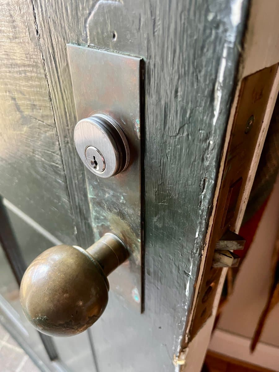A close-up photograph of a dark, weathered wooden door featuring a round brass doorknob and a new, silver-colored deadbolt lock mounted above it.