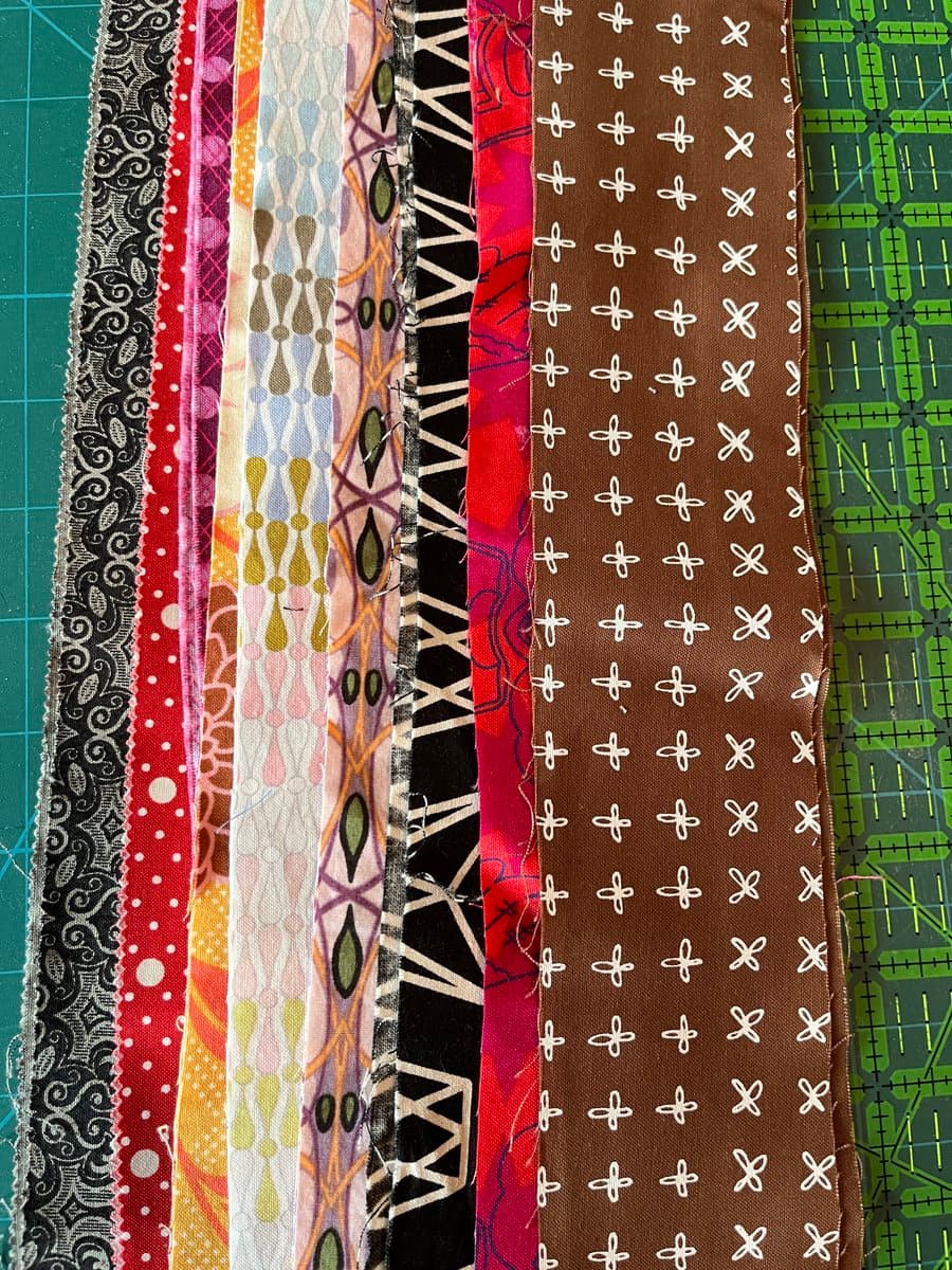 A collection of colorful fabric strips with various patterns, including lace, polka dots, and geometric designs, laid out side-by-side on a green cutting mat. The strips feature a range of colors such as black, red, pink, white, brown, and green.