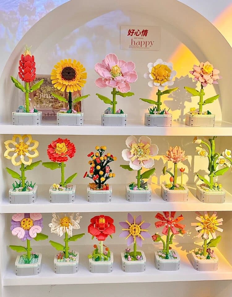 A display of colorful LEGO flower sets arranged on three white shelves, featuring various blooms like sunflowers and roses in small planters. A sign on the back wall reads '好心情 happy'.
