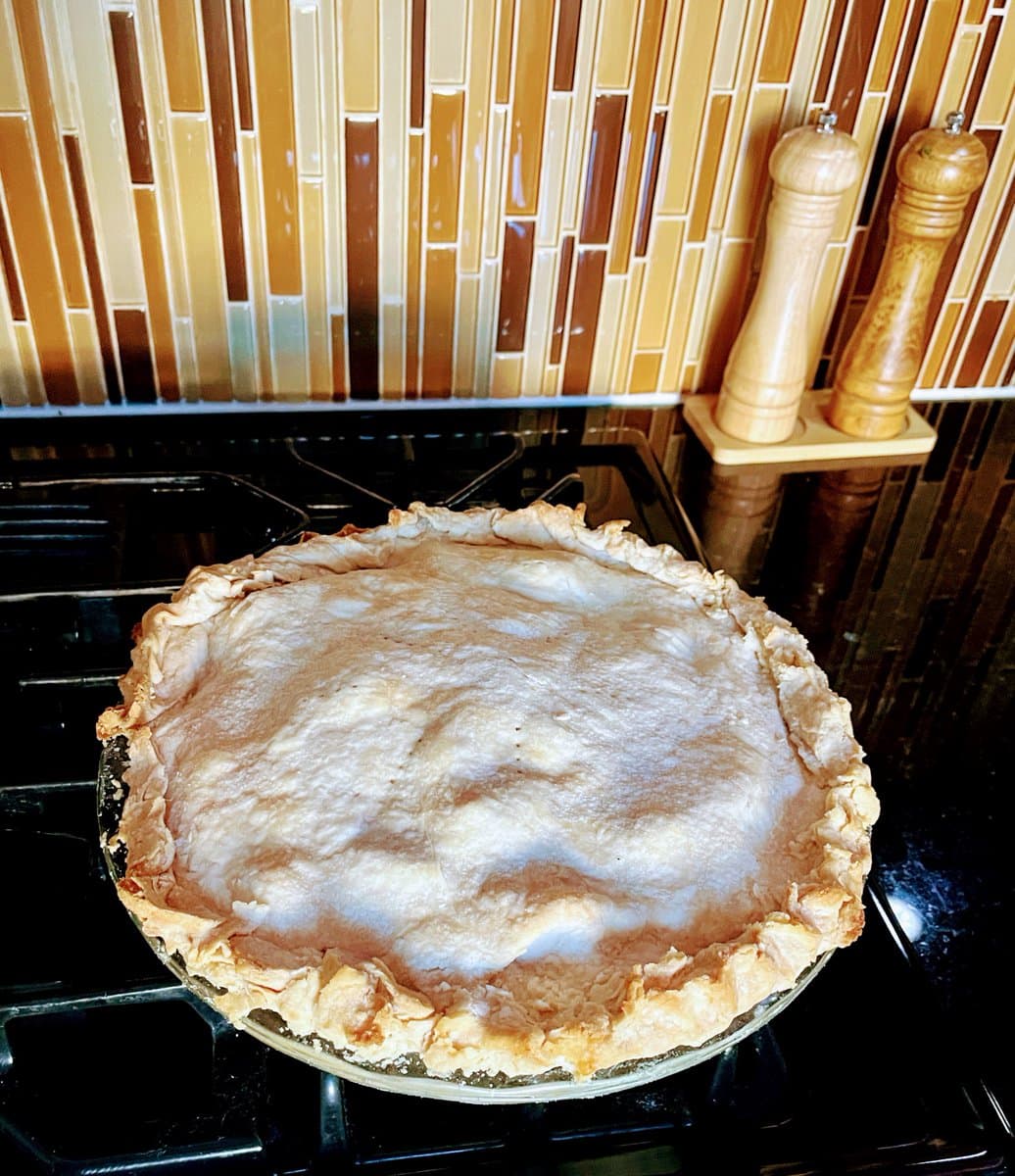 A freshly baked apple pie with a crimped crust sits on a black stovetop, set against a background of vertical amber and brown glass tiles and wooden salt and pepper grinders.