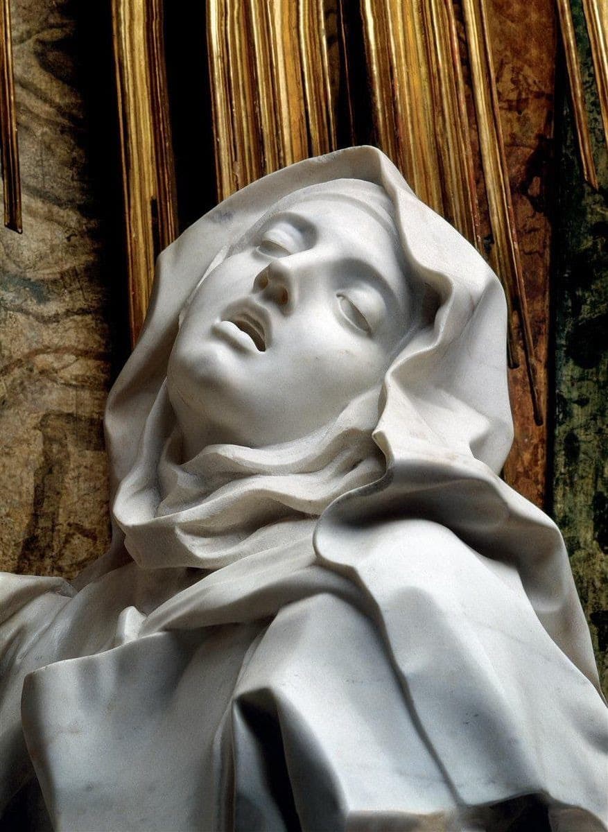 A close-up photograph of a white marble sculpture depicting a figure with a serene, upward-looking face and draped fabric, set against a background of gold and brown vertical elements.