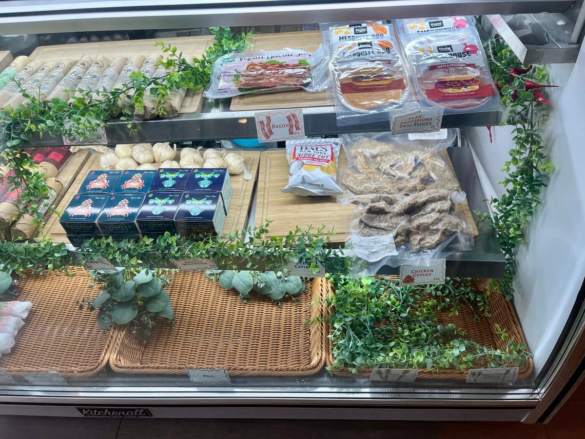 A photograph of a refrigerated display case in a vegan butcher shop, filled with plant-based meat alternatives and other products arranged on wooden shelves and wicker baskets decorated with greenery.