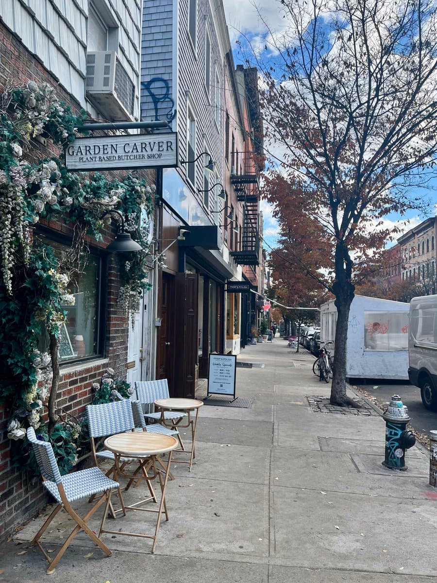 A street-level view of a storefront with a hanging sign reading "GARDEN CARVER PLANT BASED BUTCHER SHOP," decorated with artificial flowers and outdoor seating. The sidewalk extends down the block past a tree and a white food truck.