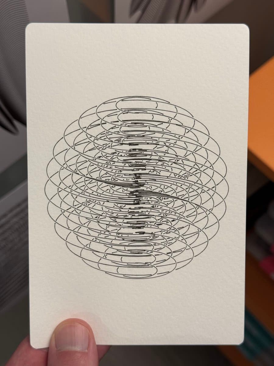A hand holds a white card featuring a black line drawing of a 3D sphere composed of stacked, overlapping rings. The background is a blurred indoor setting.
