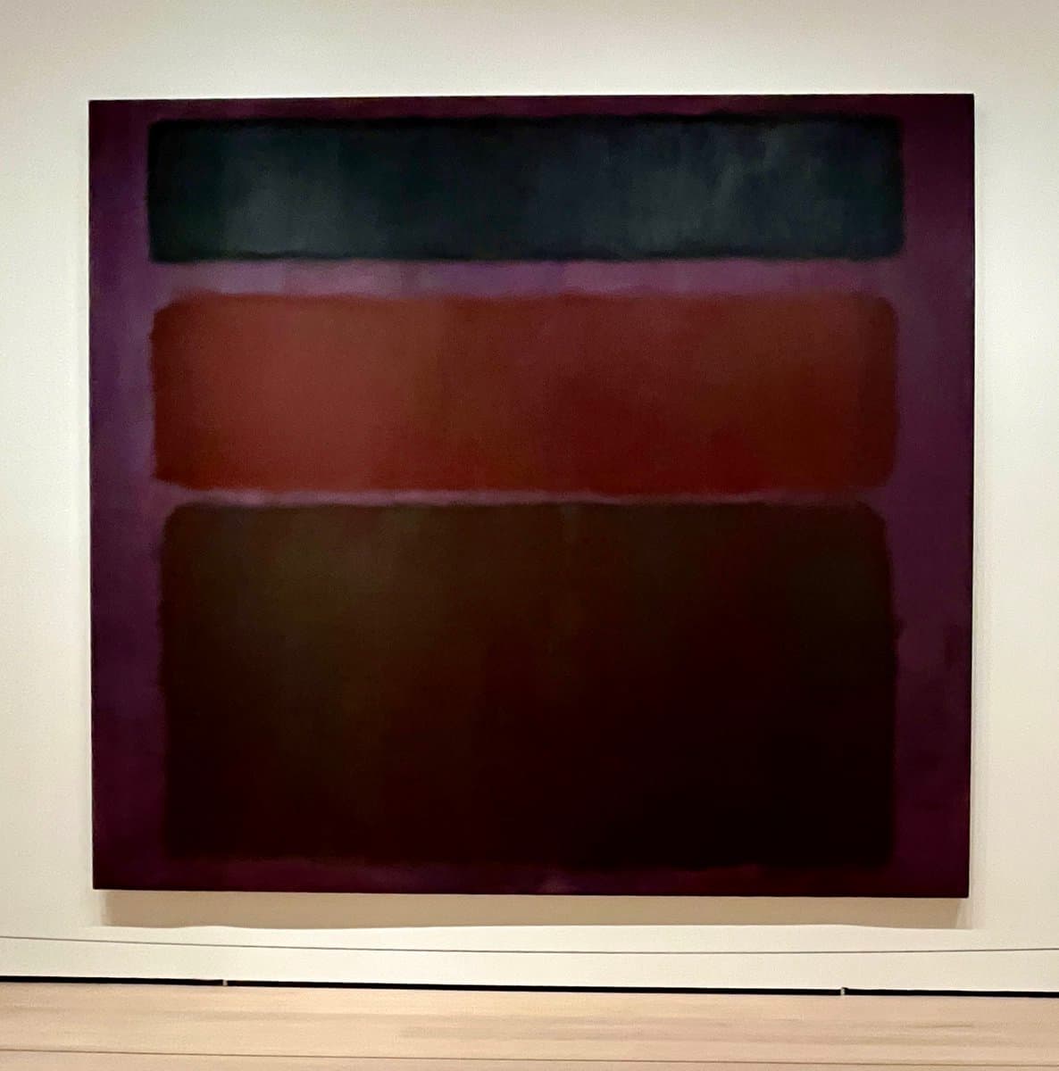 A photograph of an abstract painting hanging on a white gallery wall, featuring three horizontal bands of dark blue, reddish-brown, and dark brown against a purple background. The artwork is displayed in a museum setting with a light-colored floor visible at the bottom.